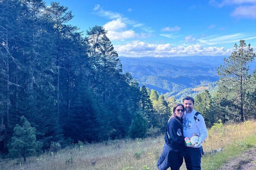 A couple standing on a forested hill with a view of distant mountains. 3