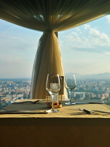 A table with two wine glasses and a bottle, set against a cityscape view through a window with a draped curtain. 2