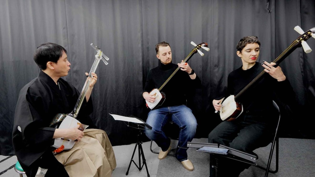Three individuals seated, each holding a stringed instrument, against a dark backdrop. 3