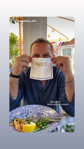 A person holding a white paper bag with 'VINECO' printed on it, sitting at a table with a blue and white patterned plate containing food. 5