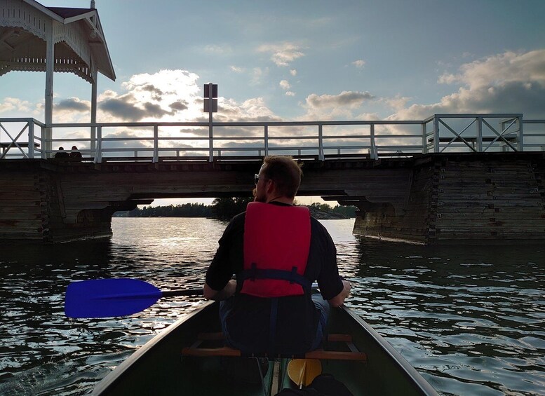 A person in a canoe on a river, wearing a life jacket, under a bridge with a railing. 2