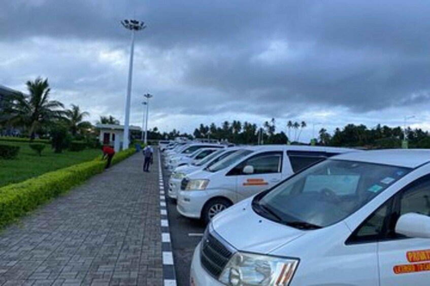 A row of parked cars along a paved walkway with palm trees and a cloudy sky in the background. 1
