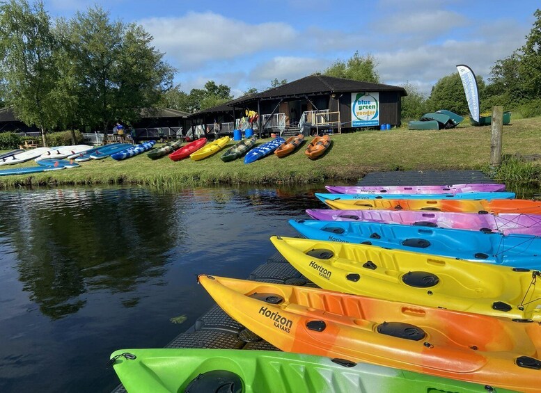 A row of colorful kayaks lined up by a calm river, with a building and trees in the background. 3