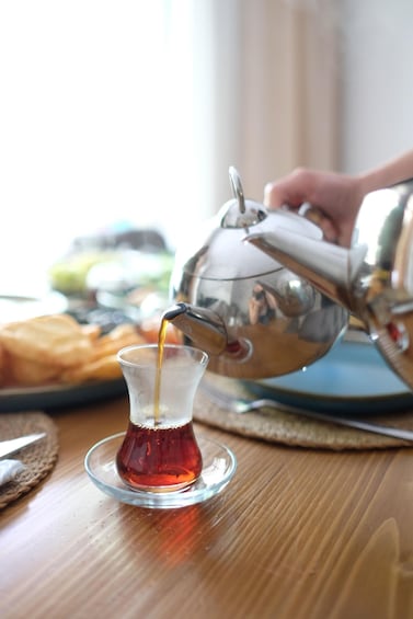A hand pouring tea from a silver teapot into a glass cup on a wooden table. 2