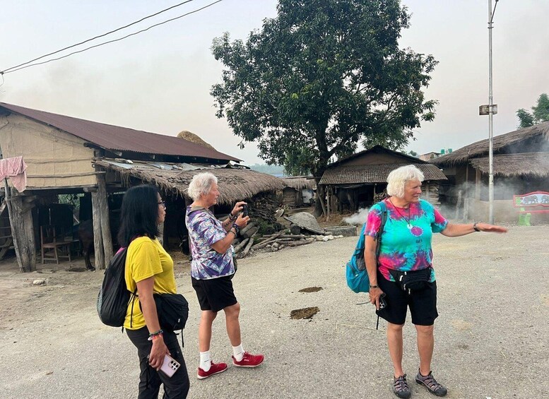 Three people standing in a rural area with thatched-roof buildings and a large tree in the background. 2