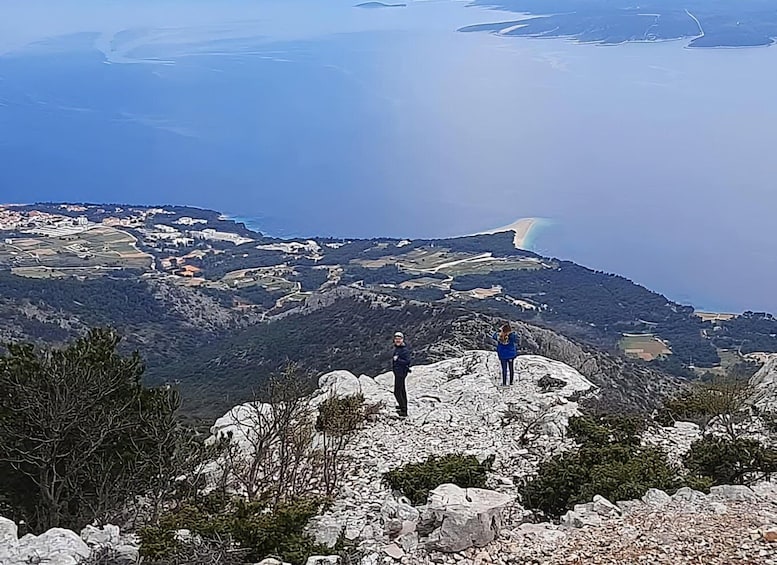 Two people standing on a rocky outcrop overlooking a coastal landscape with a beach and a town in the distance. 1