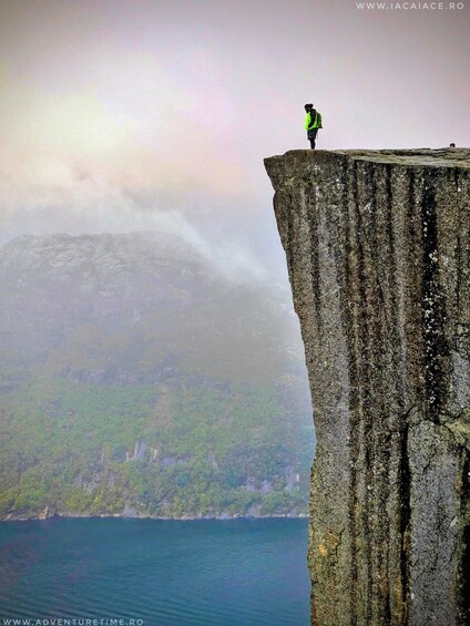 A person standing on a cliff edge with a mountainous landscape and a body of water in the background. 3