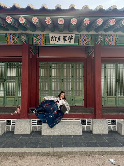 A person in traditional attire sitting on a bench in front of a building with green doors and a sign with Chinese characters. 3