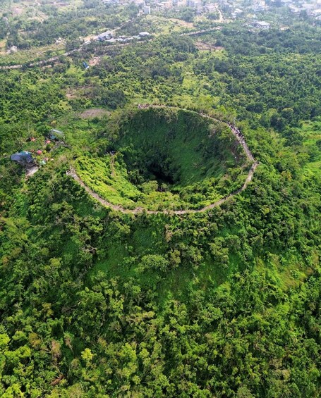 A large circular depression surrounded by dense forest, with a few buildings and a road visible in the background. 1