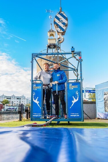 Two people on a bungee platform with a blue and white striped safety harness, a clear blue sky, and a building in the background. 1
