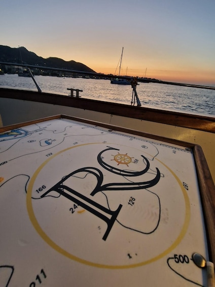 A nautical chart with a compass rose and route numbers, viewed from a boat at sunset with a marina and hills in the background. 3