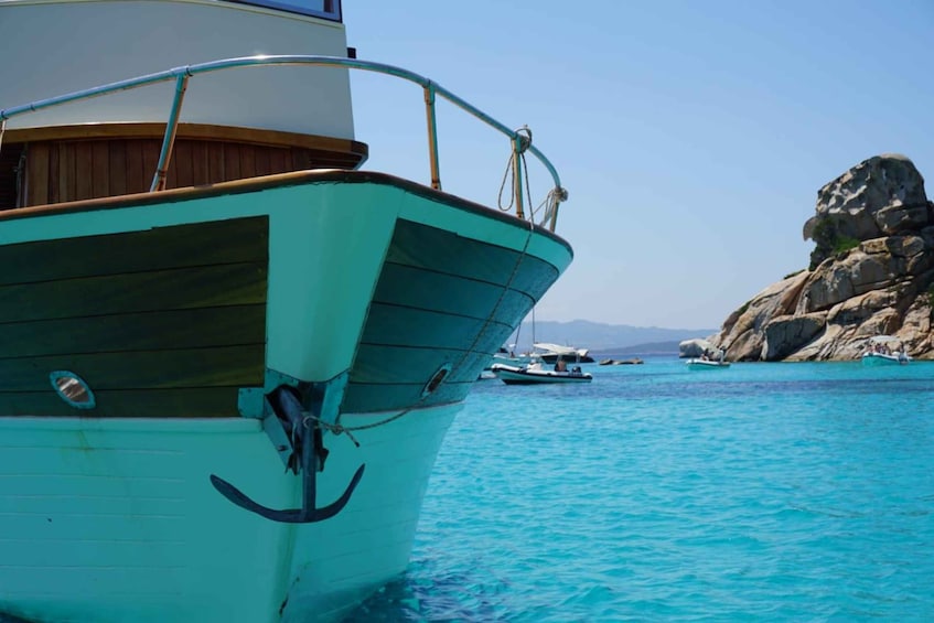 A turquoise boat anchored near rocky coastline with clear blue waters and distant mountains. 1