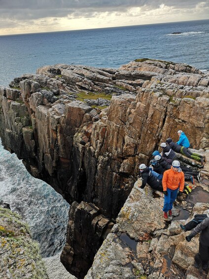 A group of people on rocky terrain near the ocean, some wearing helmets and harnesses. 1