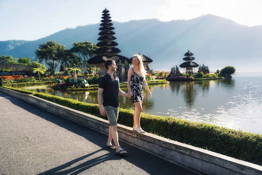 A couple walking along a lakeside path with pagodas and mountains in the background. 1