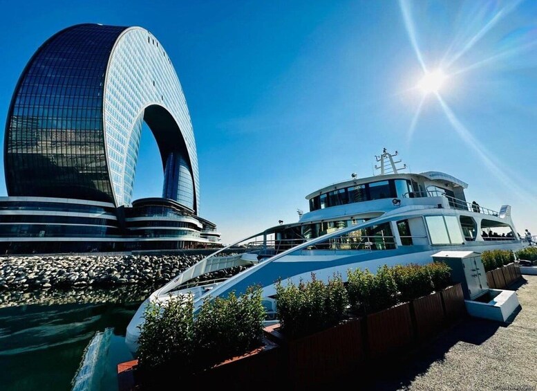 A modern glass building with a curved entrance, a white boat docked nearby, and a clear blue sky. 1