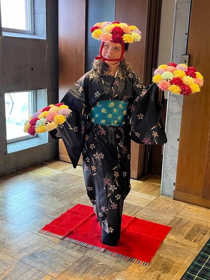Una persona in kimono giapponese tradizionale con motivi floreali, che tiene un bouquet di fiori, in piedi su un tappeto rosso in una stanza con pavimento in legno e un'ampia finestra. 2
