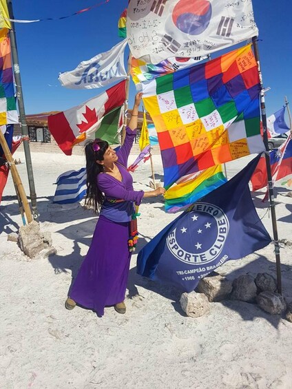 Una persona sostiene una bandera colorida con un emblema azul, de pie en una playa arenosa con varias otras banderas y un cielo azul despejado. 2