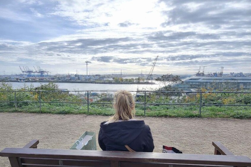 A person sitting on a bench overlooking a waterfront with industrial structures and cranes. 5