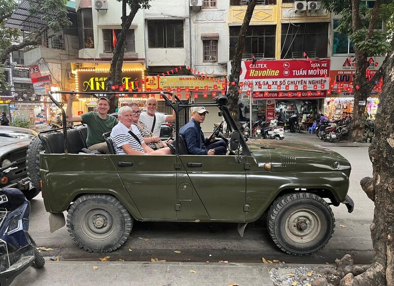 A green jeep with passengers, parked on a busy street with shops and signs in the background. 2