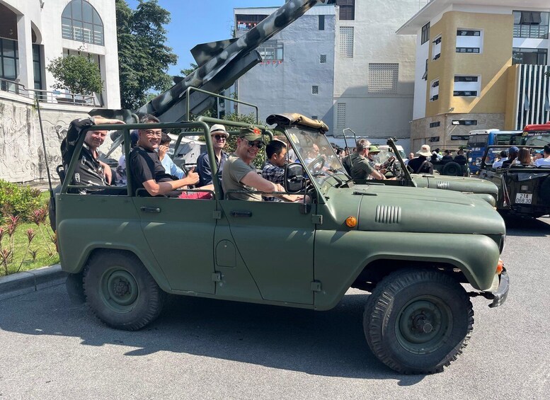 A military vehicle with a mounted cannon, passengers, and urban buildings in the background. 1