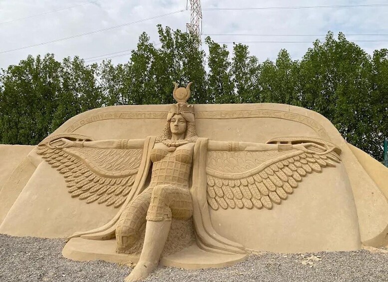 A large sand sculpture of a figure with wings, set against a backdrop of trees and a power line. 5