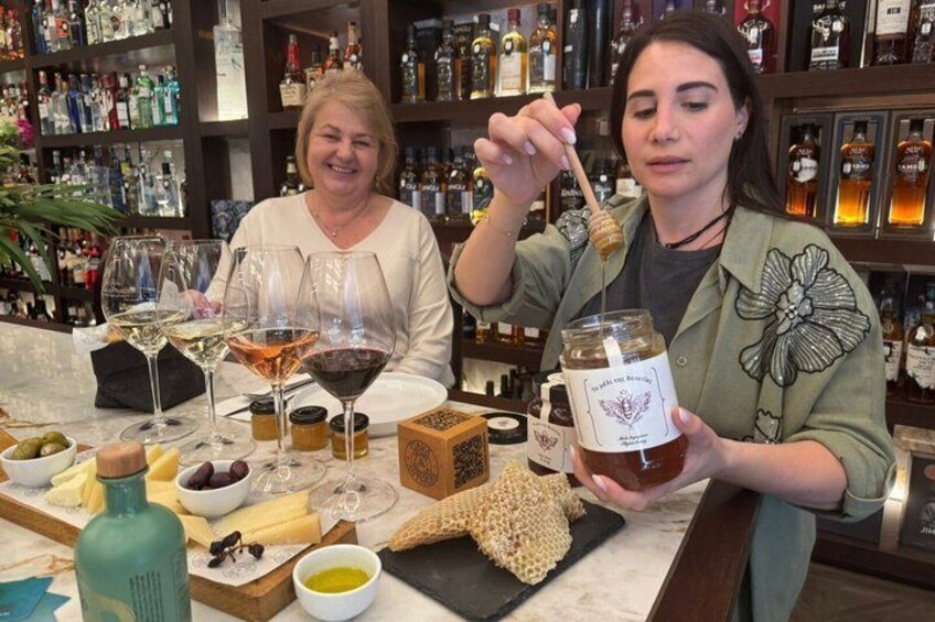Two people at a bar with wine glasses, honey jars, and a variety of bottles in the background. 1