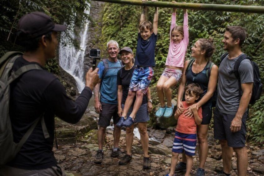 Un gruppo di persone penzolano da una barra orizzontale in una zona boschiva, con una cascata sullo sfondo. 1