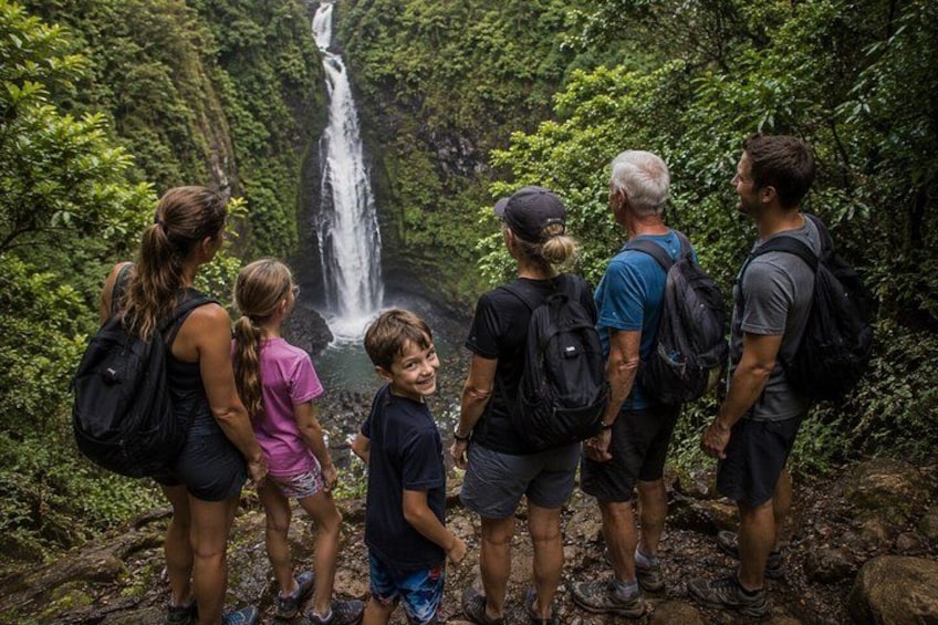A group of people standing on a rocky path, looking at a waterfall in a forested area. 4