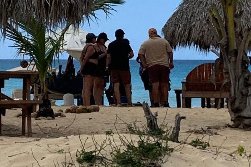 Une plage où l’on voit des gens debout et en train de marcher, un banc en bois et une structure recouverte d’un toit de chaume. 3