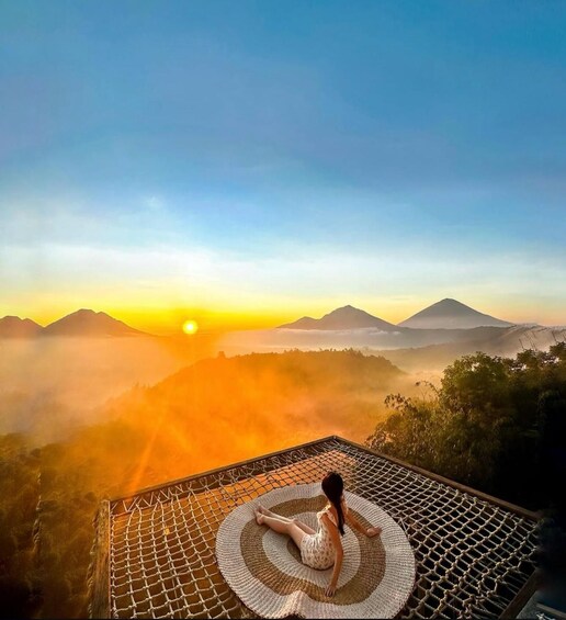 A person sitting on a woven mat on a rooftop, overlooking a mountainous landscape during sunrise. 1