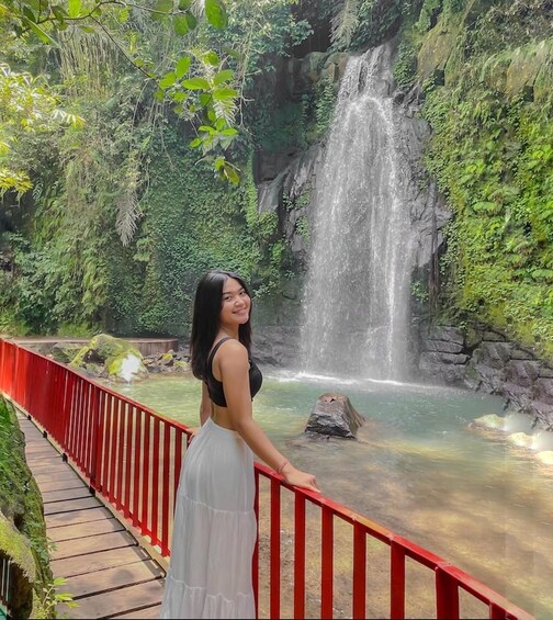 A woman in a white dress standing on a red bridge overlooking a waterfall. 2