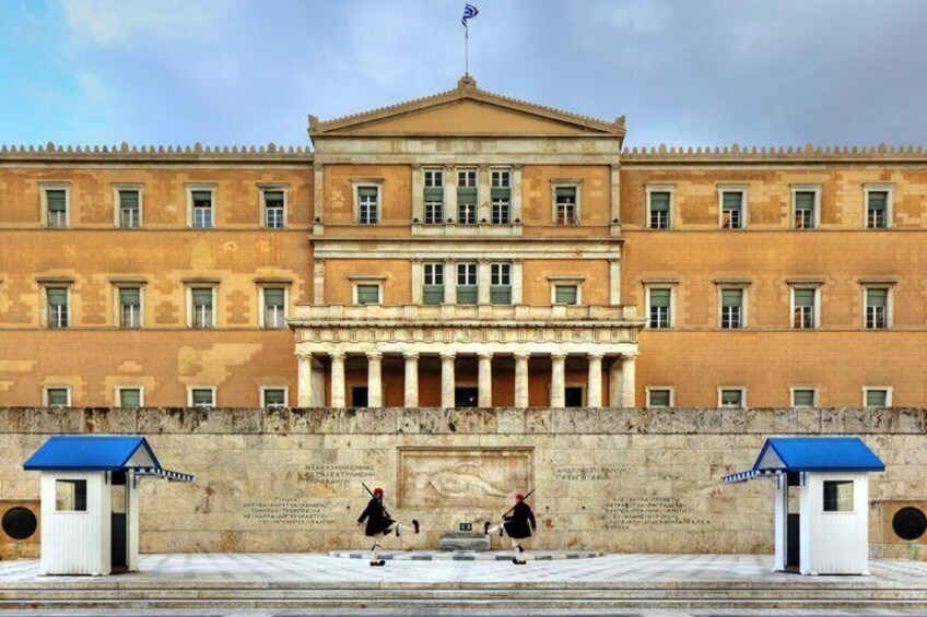A classical building with columns and a central entrance, flanked by two small structures with blue roofs. 2