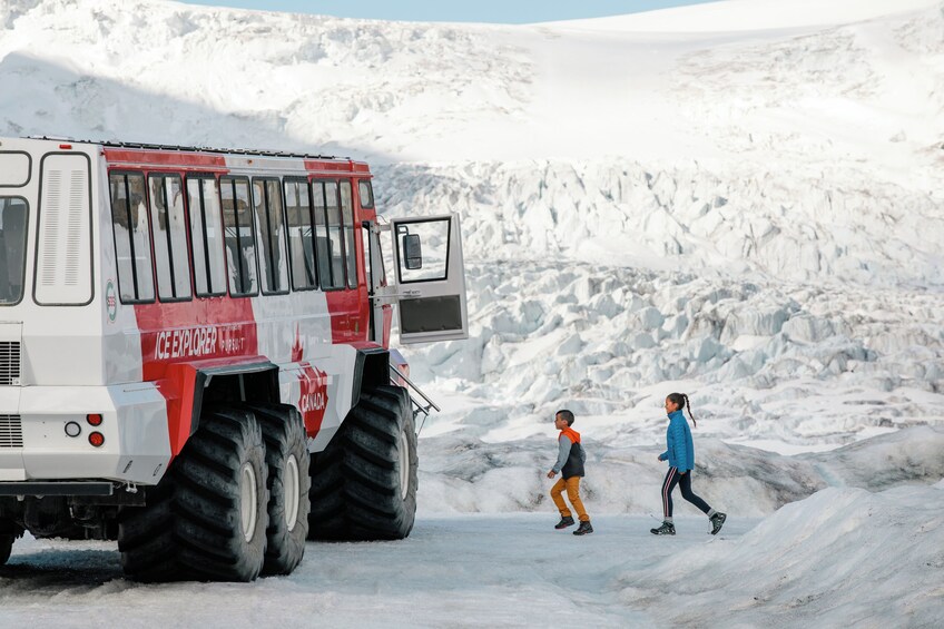 Un grande veicolo esploratore per il ghiaccio, rosso e bianco, su un terreno innevato, con due persone che camminano nelle vicinanze. 3
