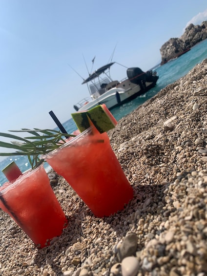 Two red drinks with straws on a pebbled beach with a boat in the background. 5
