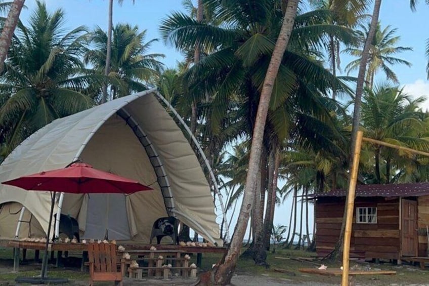 A tent with a red umbrella, a wooden table, and a small wooden structure in a tropical setting with palm trees. 3