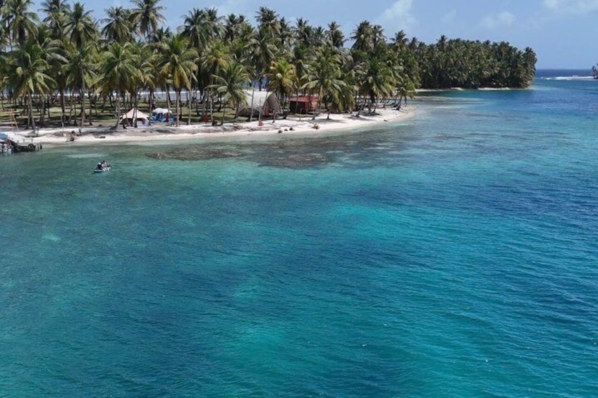 A tropical island with clear turquoise waters, palm trees, and a small beach with structures. 1