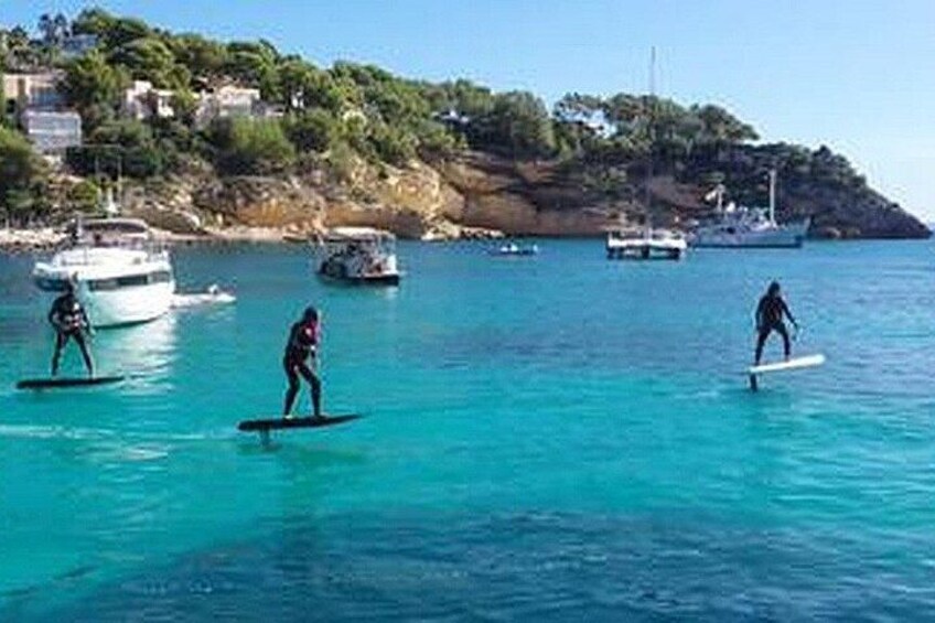 Mensen surfen op kalm turquoise water, met bootjes op de achtergrond en een rotsachtige kustlijn met gebouwen. 2