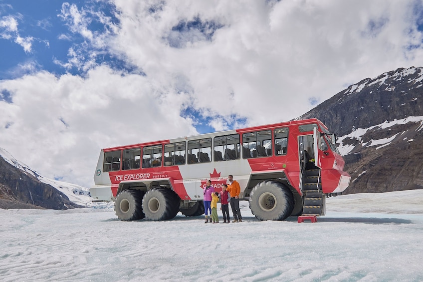 En rød og hvid turistbus med teksten 'ICE EXPLORER' og 'CANADA', parkeret på en snedækket gletsjer med bjerge i baggrunden. 1