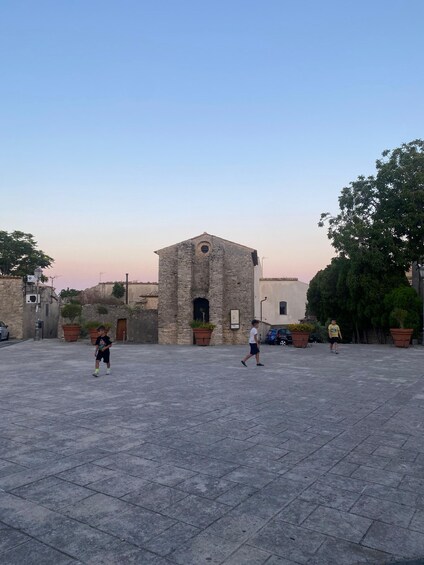 A stone building with a central archway, surrounded by potted plants and a paved square. 4