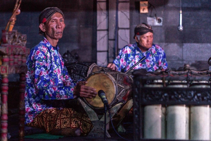 Two individuals playing drums in a room with patterned walls and traditional decorations. 5