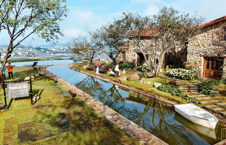 A tranquil riverside garden with stone buildings, a boat, and a sign for a coffee shop. 5