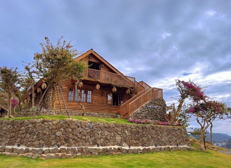 A wooden cabin with a stone foundation, surrounded by lush greenery and flowering trees. 2