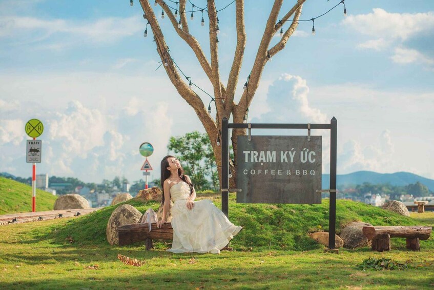 A woman in a white dress sitting on a log near a sign that reads 'TRAM KY ÚC COFFEE & BBQ.' 1
