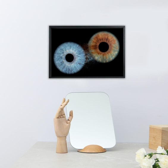 A framed artwork of two eyes on a wall, a wooden hand sculpture, and a white-framed mirror on a table. 5