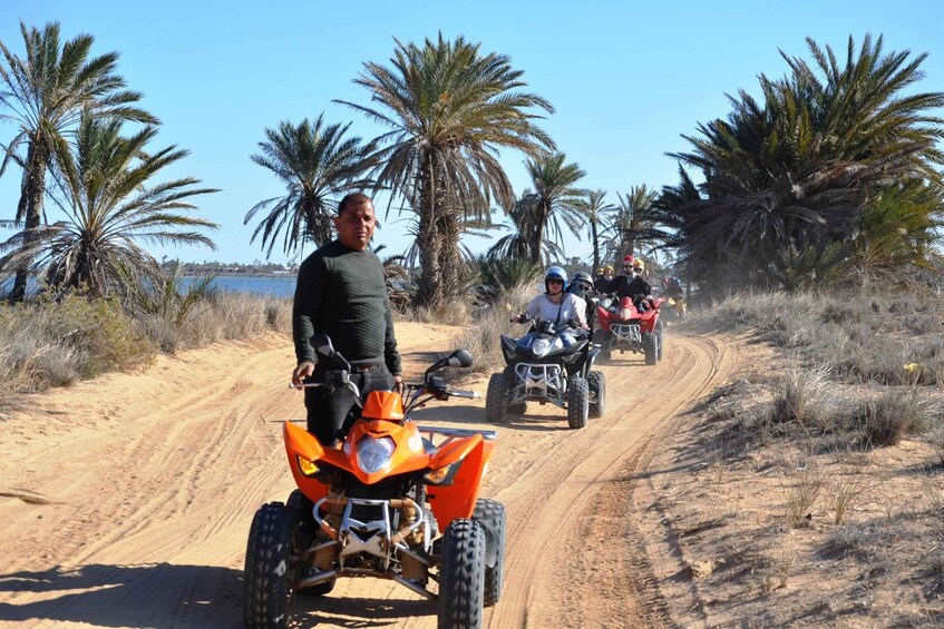 A group of people on ATVs on a sandy path with palm trees and a body of water in the background. 1