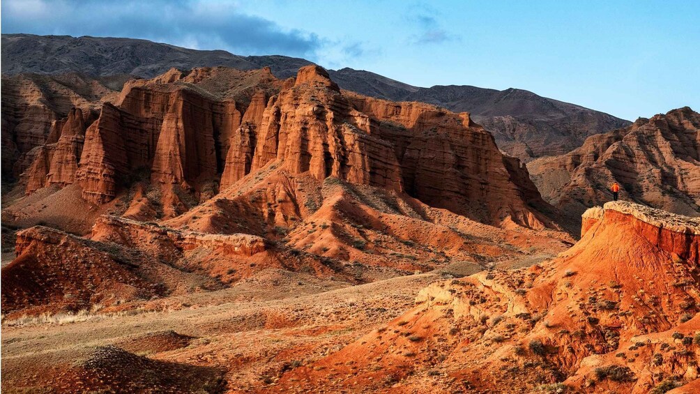 A rugged desert landscape with prominent red rock formations and a person standing on a cliff edge. 3