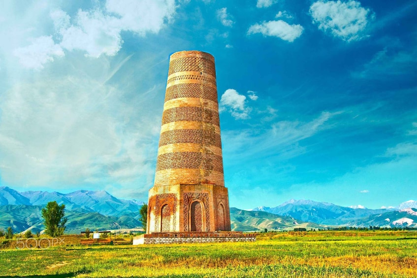 A tall, cylindrical tower with intricate brickwork, set against a mountainous backdrop under a clear sky. 1