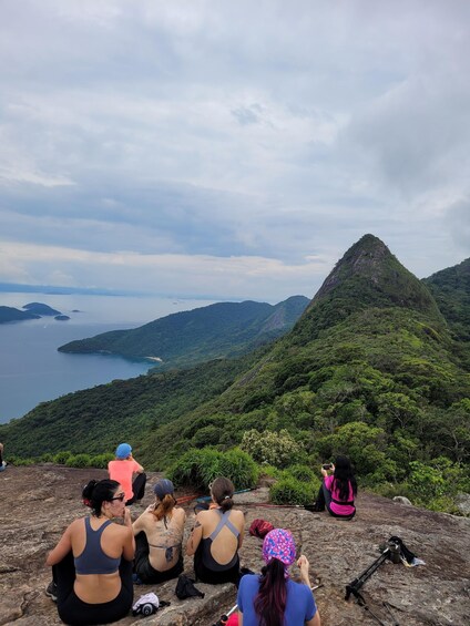 A group of people sitting on a rocky outcrop overlooking a mountainous landscape with a body of water in the distance. 1