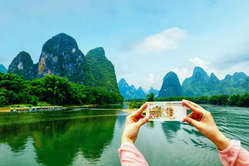 A person holding a 20 yuan note in front of a river with boats and limestone karsts in the background. 1
