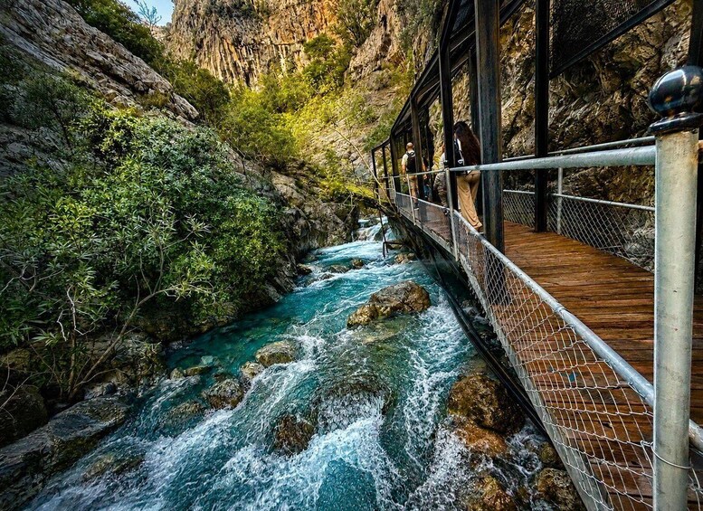 A wooden bridge over a fast-flowing river with rocky cliffs on one side and dense greenery on the other. 1